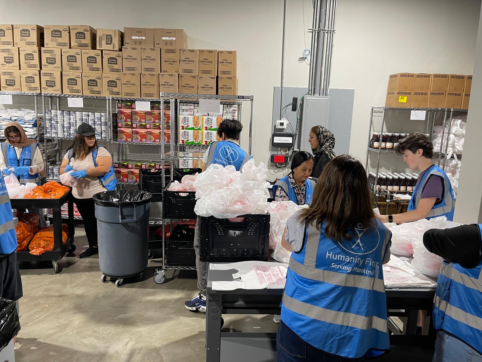 People wearing blue humanity first vests pack food at different stations in a warehouse that is packed with food items on shelves and stacked boxes.