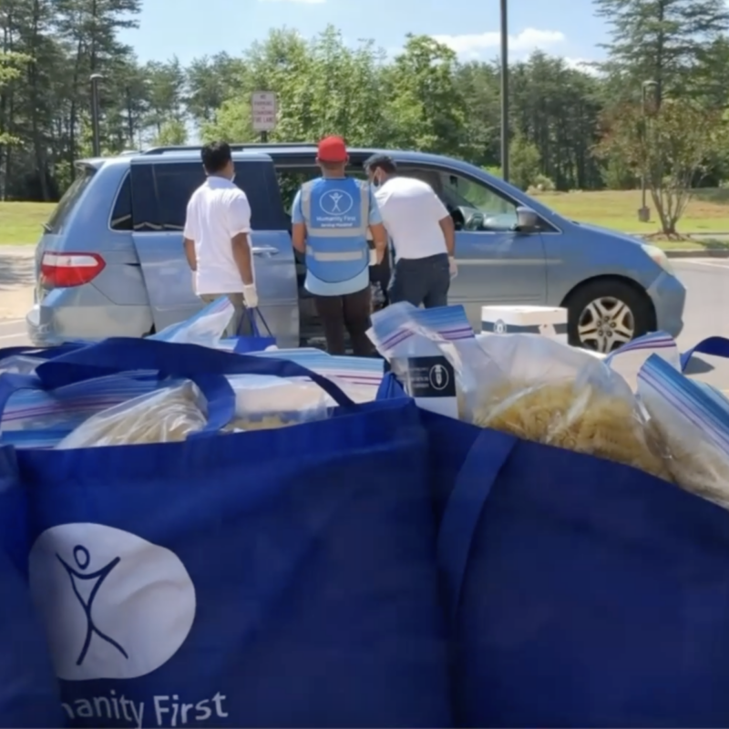 Three people put supplies into a minivan and in the foreground are blue humanity first bags packed with food staples.