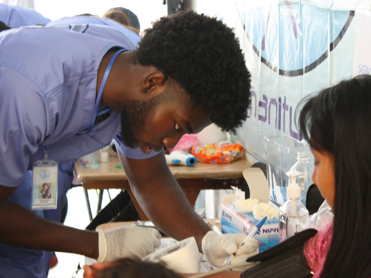 A male university student wearing blue medical scrubs looks at readings on a blood pressure device next to a patient wearing a blood pressure cuff.