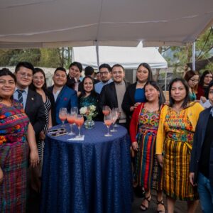 A dozen people pose around a banquet table. Some wear traditional Guatemalan clothing and others are in suits or party dresses.
