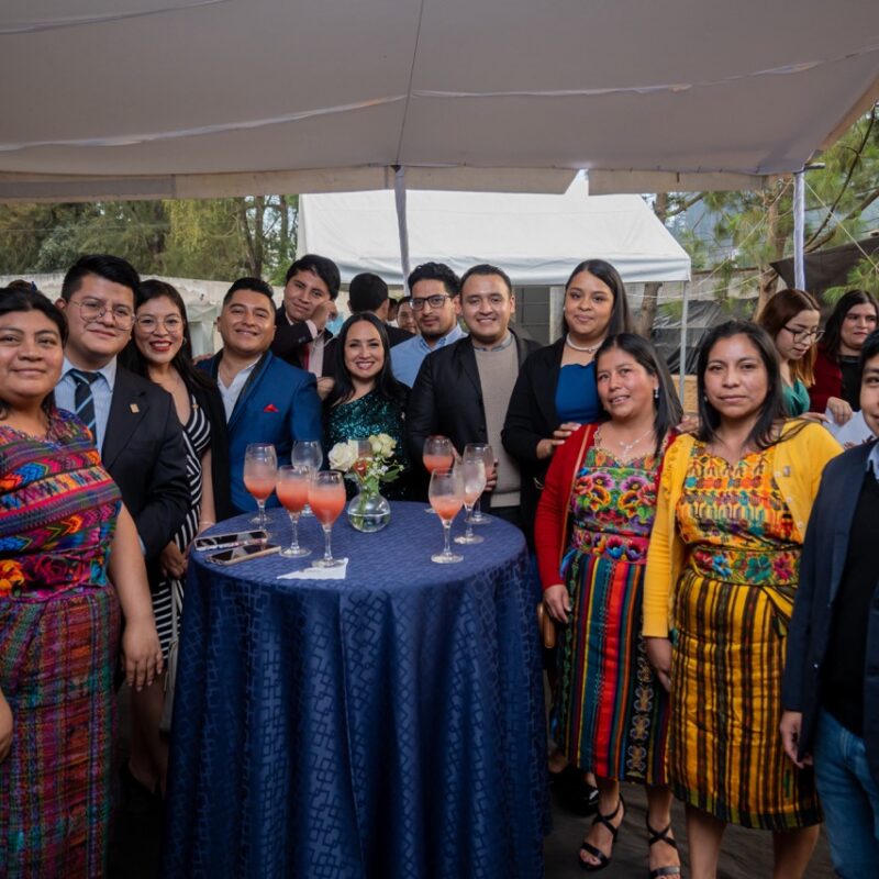 A dozen people pose around a banquet table. Some wear traditional Guatemalan clothing and others are in suits or party dresses.