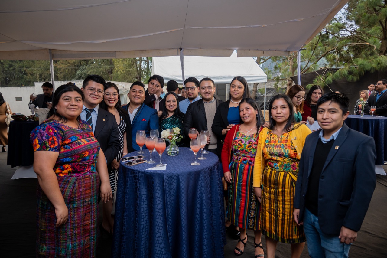 A dozen people pose around a banquet table. Some wear traditional Guatemalan clothing and others are in suits or party dresses.