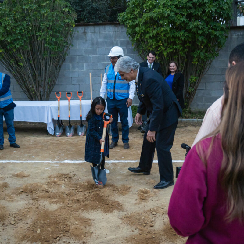 Young girl uses an adult shovel to dig into the ground at the ceremony next to a man in a suit encouraging her.