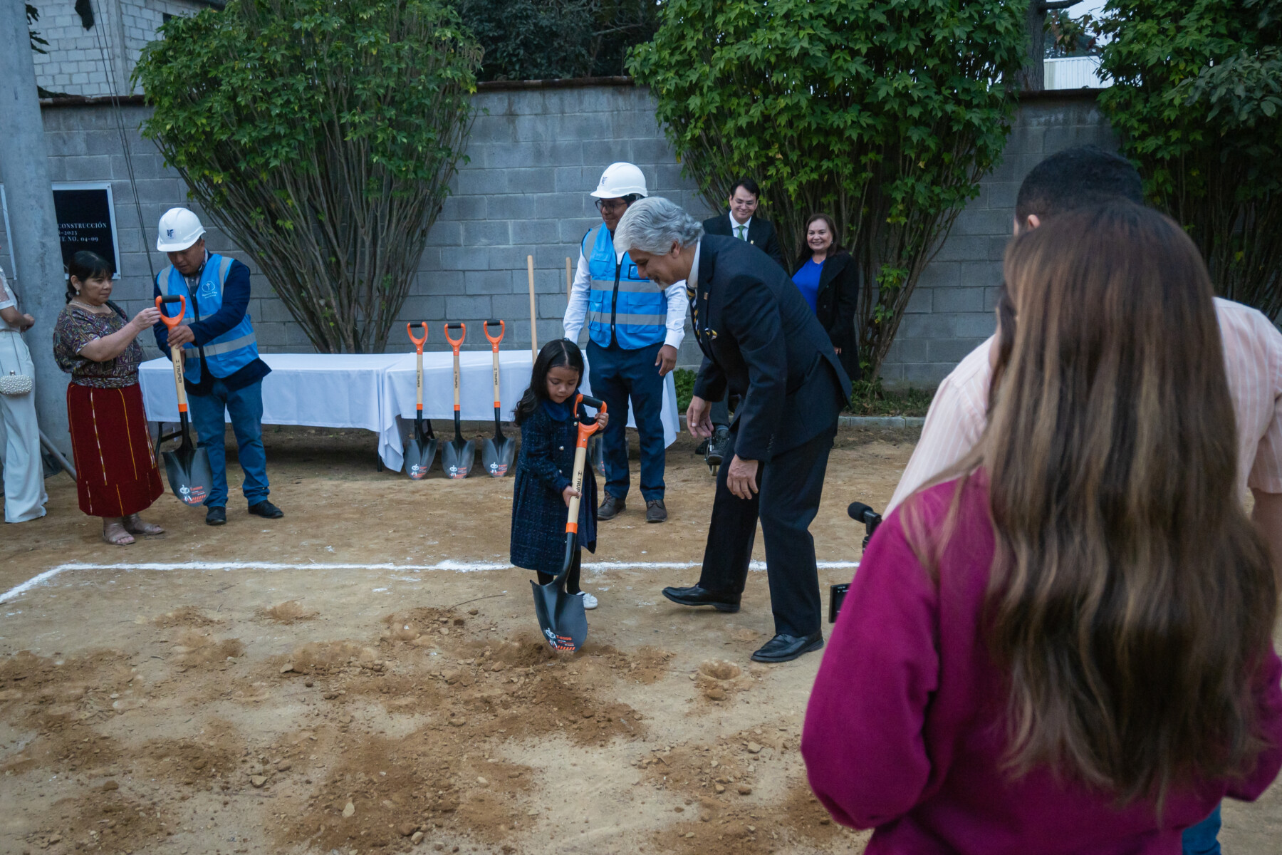 Young girl uses an adult shovel to dig into the ground at the ceremony next to a man in a suit encouraging her.