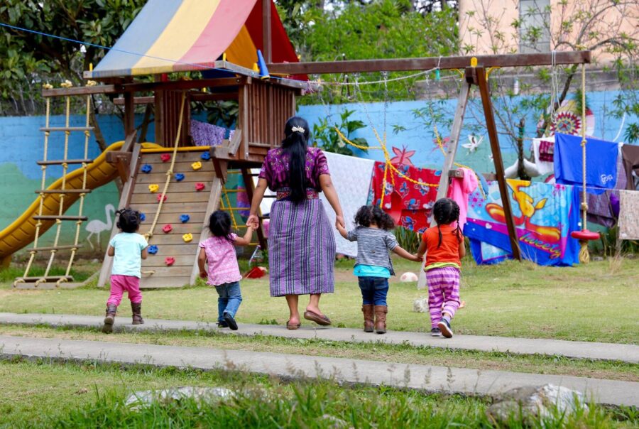 Small children walk with a women toward a playground set in the background of the image. Two of the children hold the woman's hands, a third holds the hand of another child, and the fourth walks alongside them.