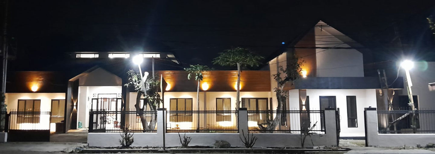A white and grey one-story building with a few small palm trees in front is lit at night with white and yellow lighting.