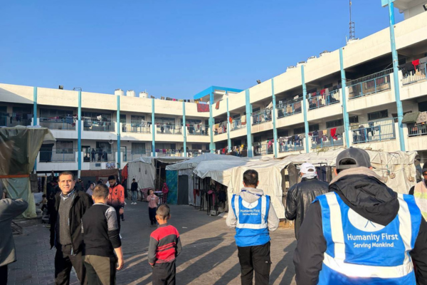 Two Humanity First volunteers with blue vests stand in a space in front of two-story building with open air hallways and railings. In the space are makeshift tents and people standing or walking through.