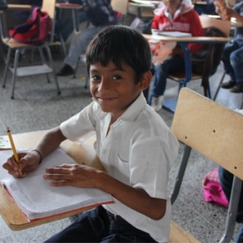 A young boy wearing a short sleeve white collared shirt sits at a wooden desk and chair and is writing with a pencil into a notebook.