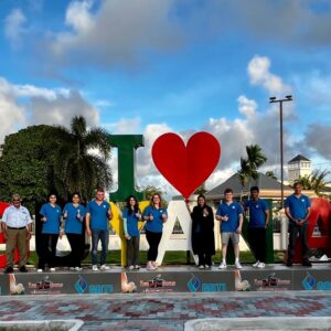 Students from the Gift of Health medical mission wear blue Humanity First polo shirts and stand in a line in front of a large sculpture that reads I heart Guyana.