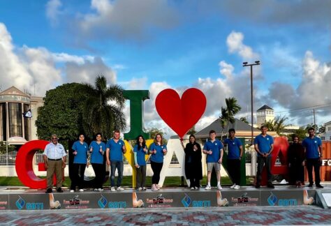 Students from the Gift of Health medical mission wear blue Humanity First polo shirts and stand in a line in front of a large sculpture that reads I heart Guyana.