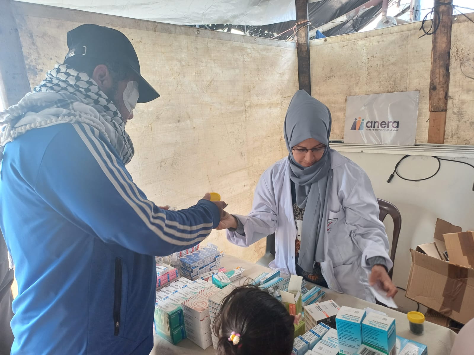 A woman in a white coat and headscarf hands a bottle to a man with a patch on his face. She stands behind a table with boxes and bottles of medicine on top under a makeshift tent structure.