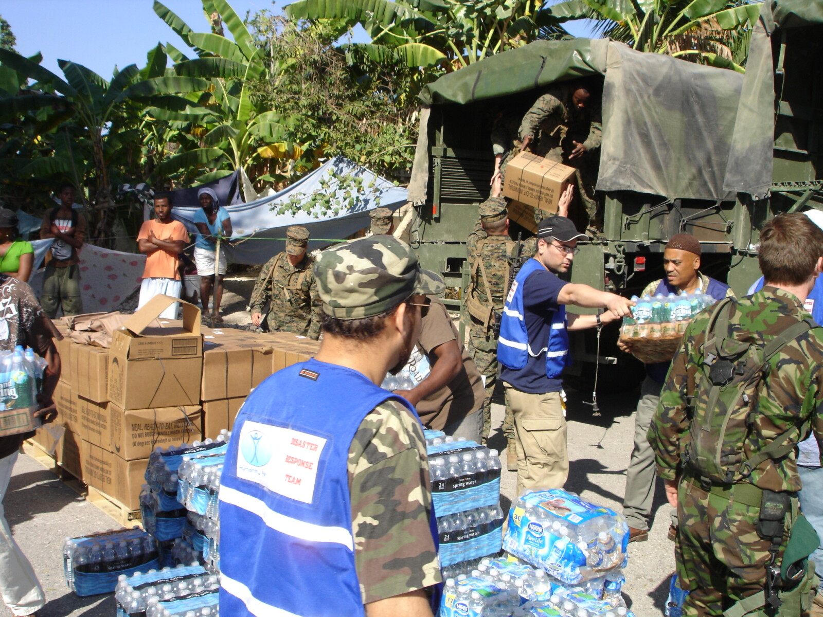 Men in fatigues and blue humanity first vests organize boxes and pallets of bottled water unloaded from a truck