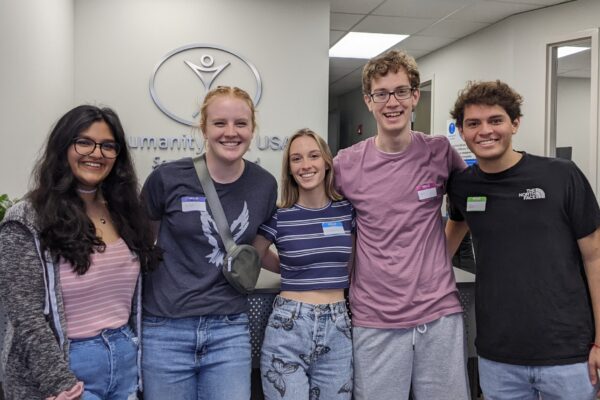 Five young professionals, three women and two men, pose together in front of a wall with the Humanity First logo on it.