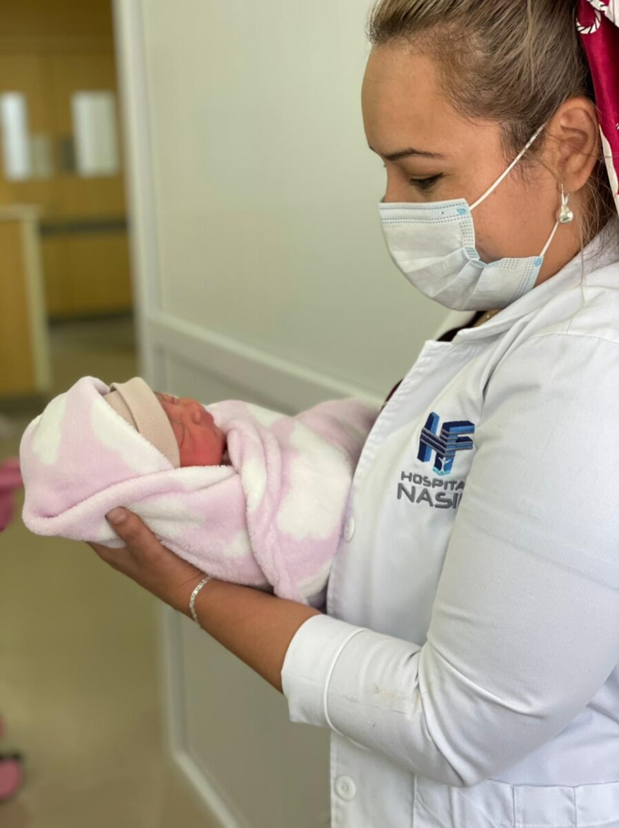 An infant is wrapped in a pink and white blanket and is held by a woman wearing a white medical coat and surgical mask who looks down at the baby in her arms.