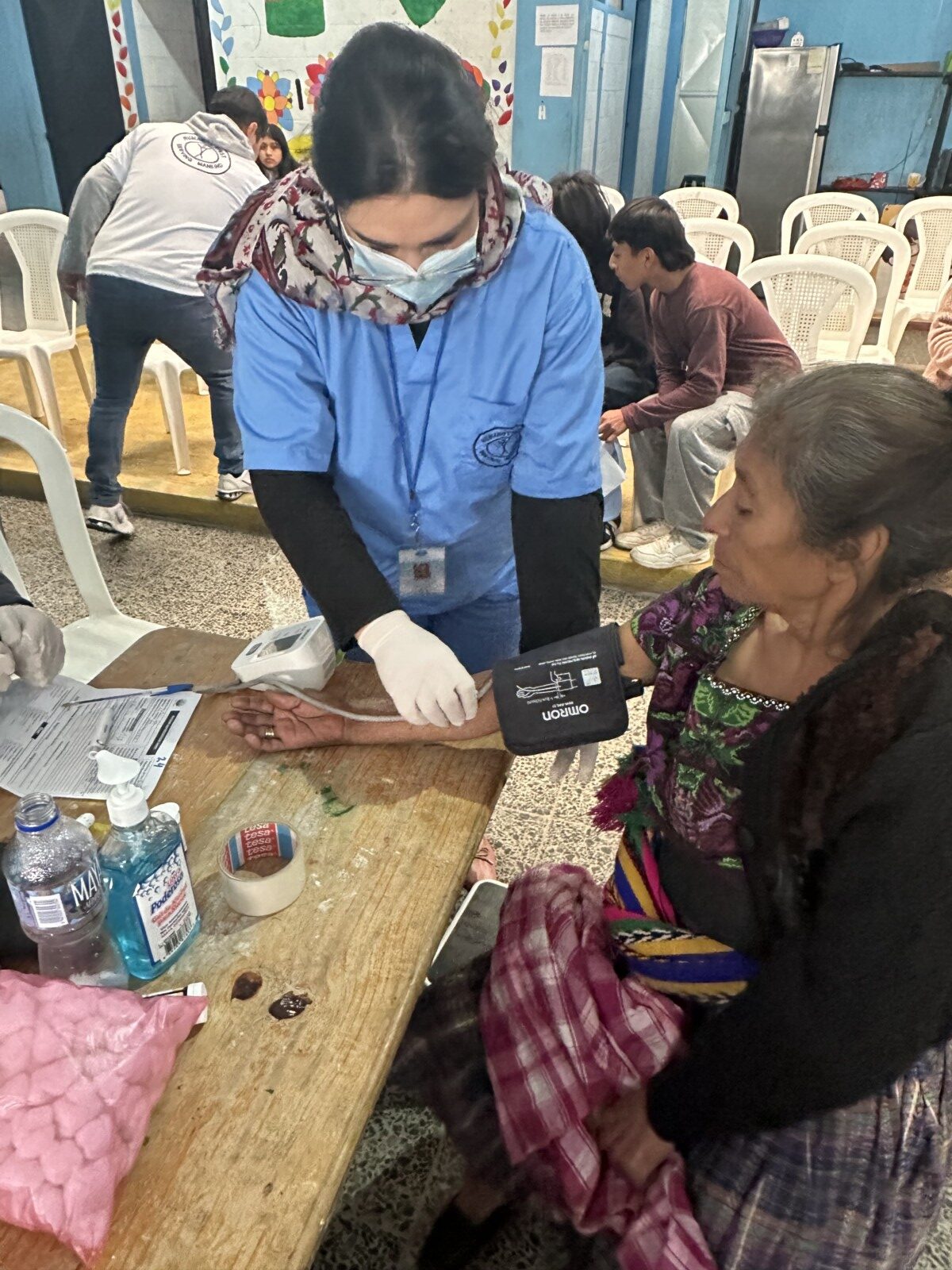 An elderly woman in traditional Guatemalan clothing gets her blood pressure taken by a female in blue Humanity First scrubs