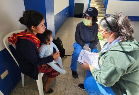 A mother with her baby speaks with two students doing a medical intake form