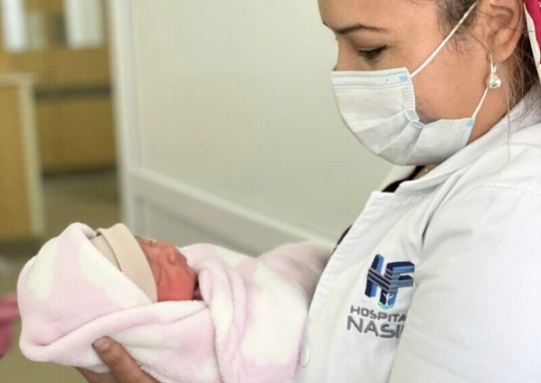 A woman wearing a white coat with the logo for HF Healthcare Hospital Nasir and a surgical mask holds and looks lovingly at a baby wrapped in a pink and white fleece blanket.