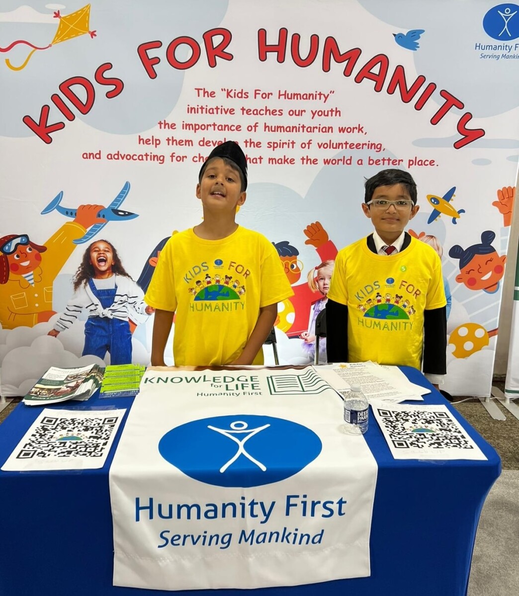 Two youth wearing yellow Kids for Humanity t-shirts stand behind a table covered with banners and fliers. Behind them is a large banner reading Kids for Humanity.