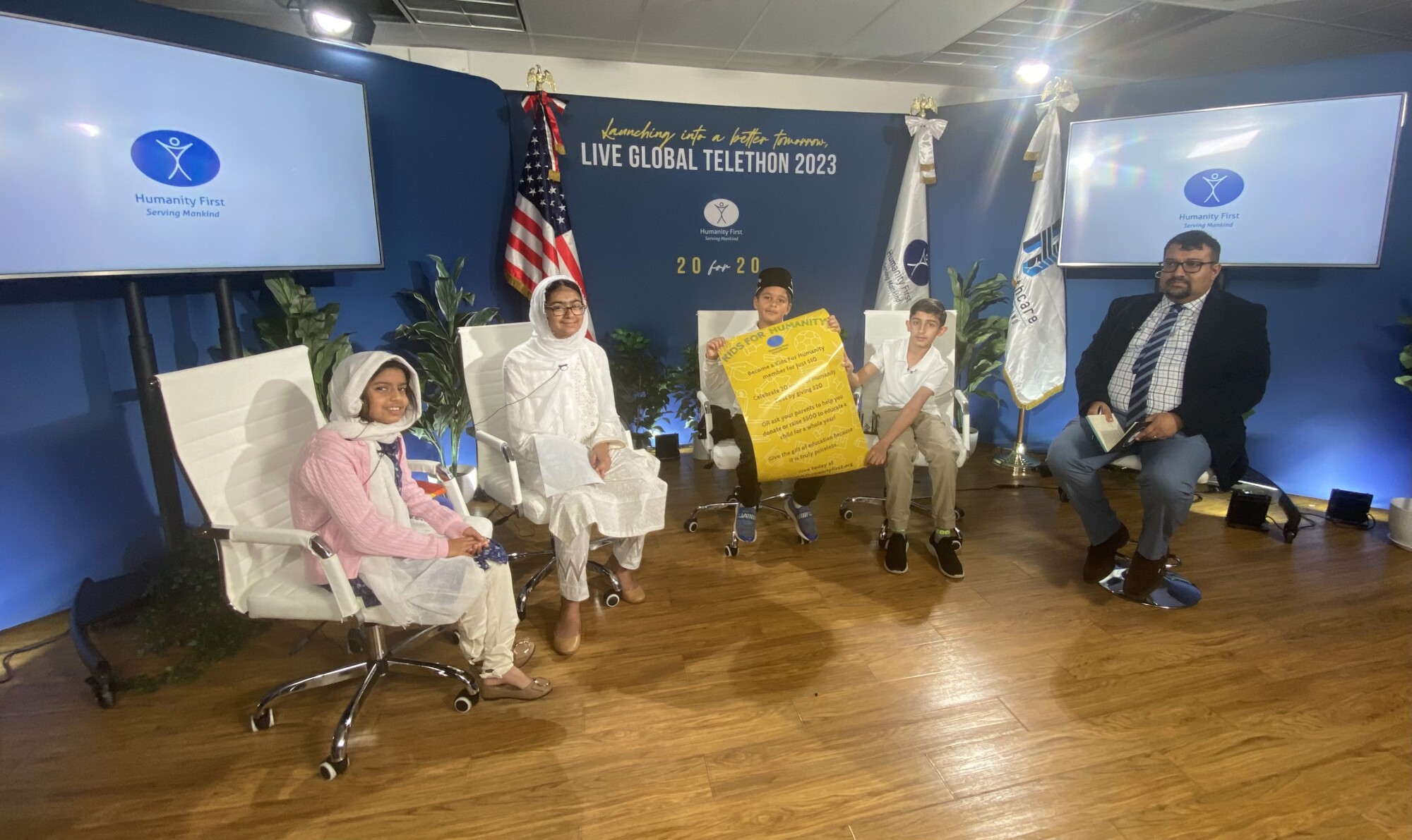 Seated left to right in front of a banner reading Live Global Telethon 2023 are a young girls wearing a pink cardigan and a white headscarf, an older girl wearing all white and a headscarf, two young boys in polo shirts holding a yellow poster, and a man in a suit.