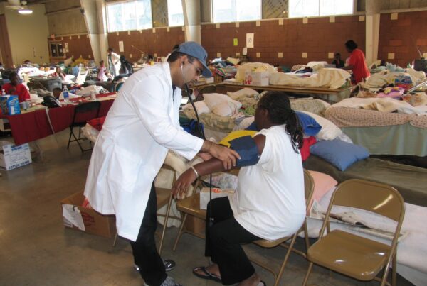 In a large open room with many tables and cots a doctor stands next to a patient seated on a folding chair and listens to her pulse while using a blood pressure cuff.