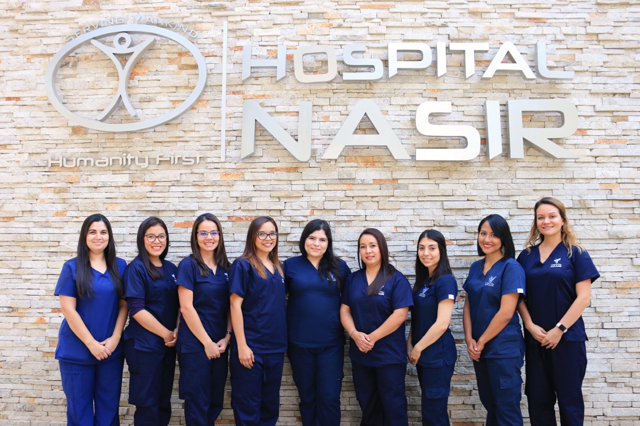 9 women in dark blue medical scrubs pose in a line in front of a tan brick wall with large sign for Hospital Nasir