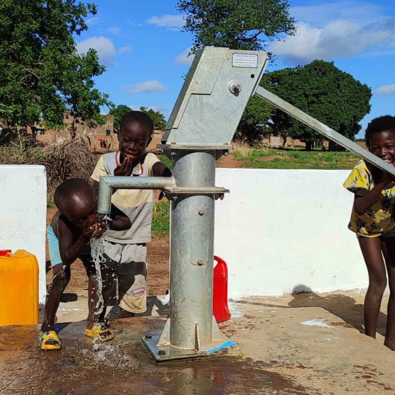 Four children laugh around a water hand pump as one pumps and another drinks from the water pouring from the spigot.