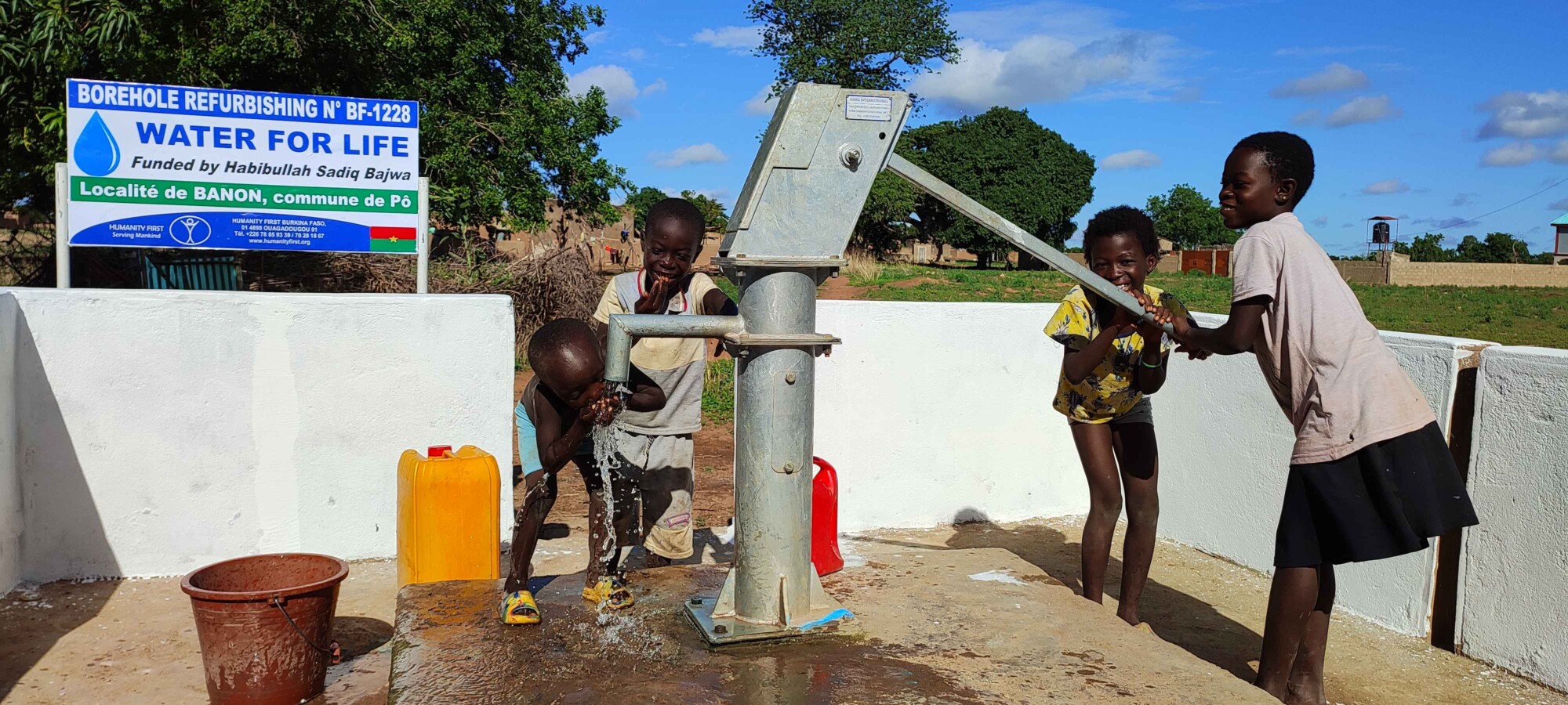 Four children laugh around a water hand pump as one pumps and another drinks from the water pouring from the spigot.