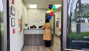 From an open doorway, the images shows an entranceway with a reception desk in front of a wall showing the Humanity First USA logo. Two women sit behind the desk which is decorated with a cluster of balloons and a houseplant. Another woman in a coat checks in at the desk.