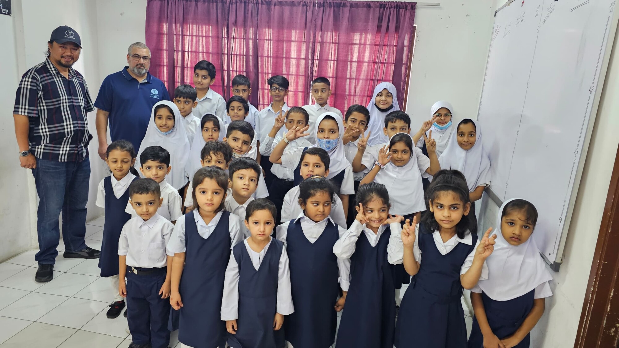 About two dozen students of multiple elementary school ages wear white and blue school uniforms and pose in a group with a member of school staff and our visiting VP of Human Development Programs.