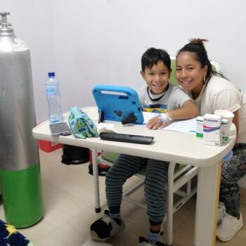 A young boy and his mom smile brightly as the boy sits at a desk in the hospital next to a large oxygen tank and his mom crouches next to him for the picture.