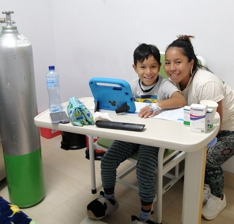 A young boy and his mom smile brightly as the boy sits at a desk in the hospital next to a large oxygen tank and his mom crouches next to him for the picture.