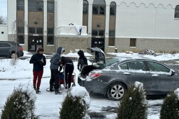 In a driveway covered in snow four people load supplies into a car with its truck popped open.