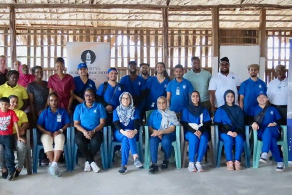 Over two dozen people pose in two rows, one standing and one sitting next to a sign reading Global Health Humanity First Guyana. They are inside a thatched building and many of them wear blue medical scrubs.