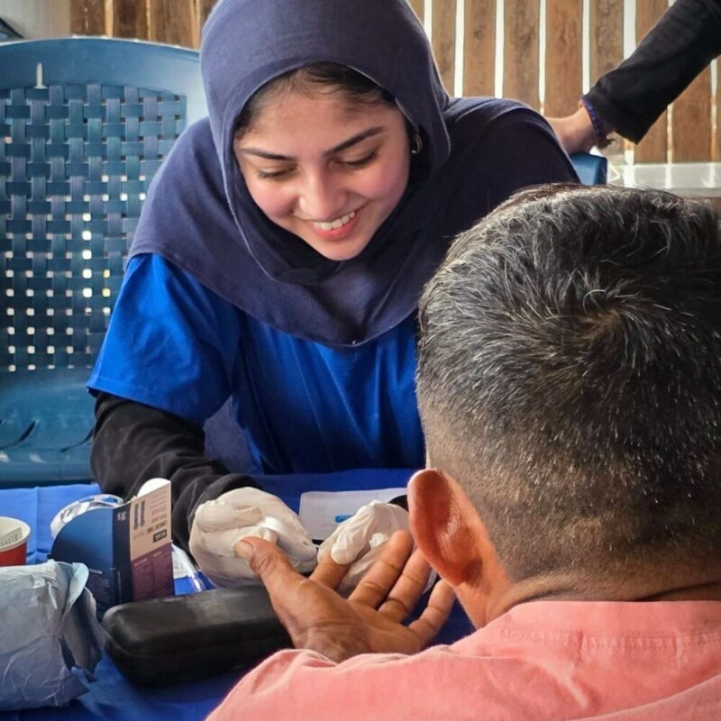 A woman wearing blue medical scrubs and a hijab takes gauze to the finger of a man with his back to the camera.