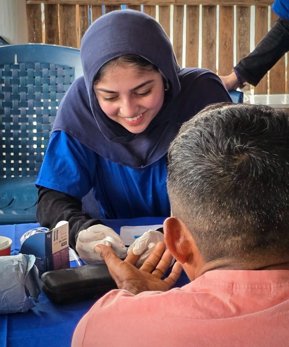 A woman wearing blue medical scrubs and a hijab takes gauze to the finger of a man with his back to the camera.
