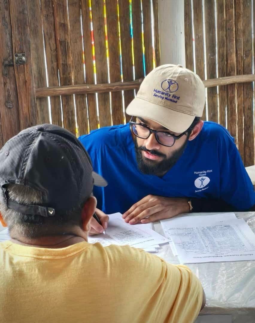 A man wearing a baseball cap and polo shirt with the logo for Humanity First looks intently at a man in a baseball cap and yellow t shirt and writes onto a paper form