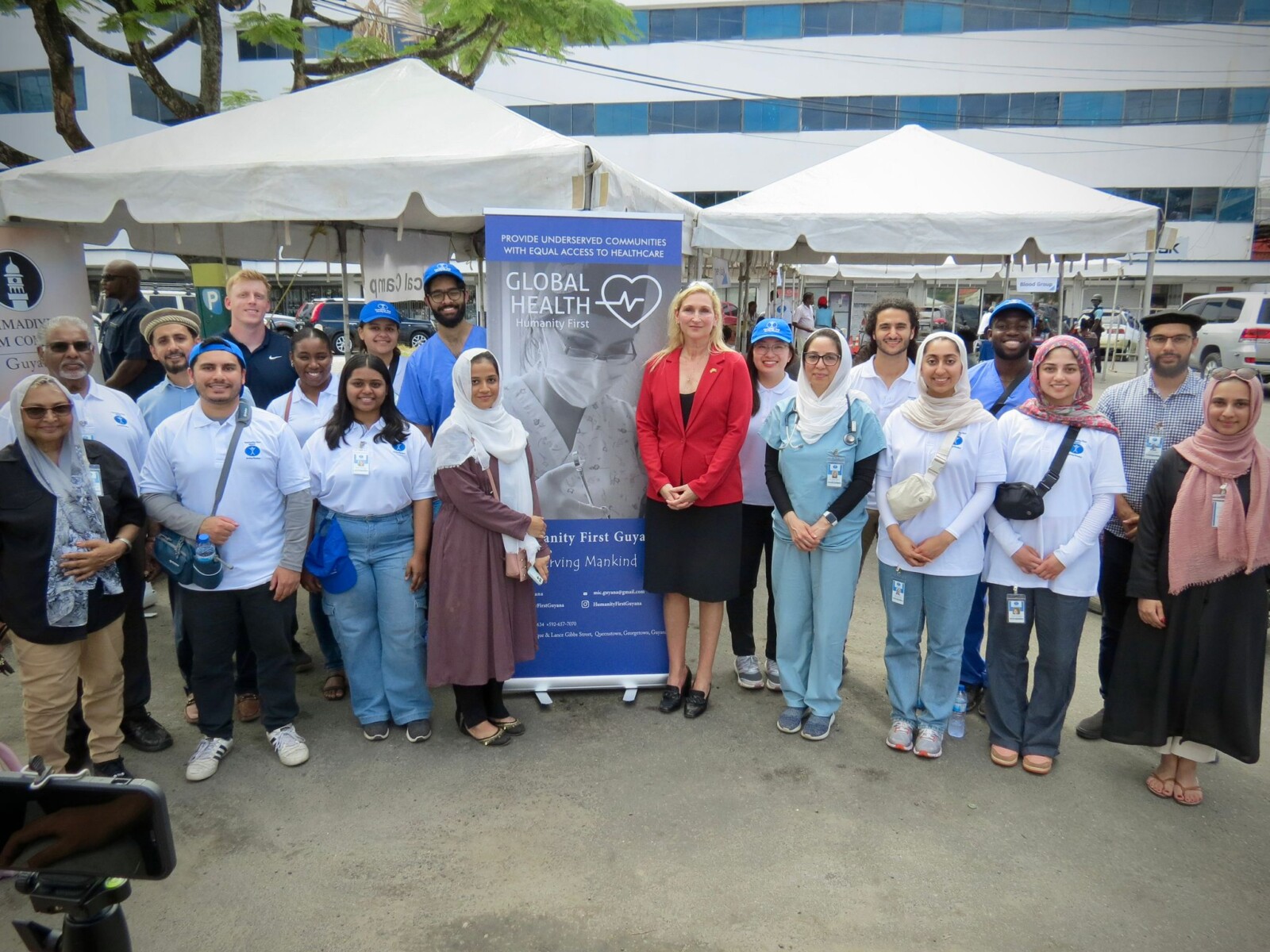 A group of people pose standing outdoors next to a sign reading Global Health. Some of the groups wears white t shirts or medical scrubs and baseball caps or hijabs. In the center is a tall blonde woman in a red blazer.