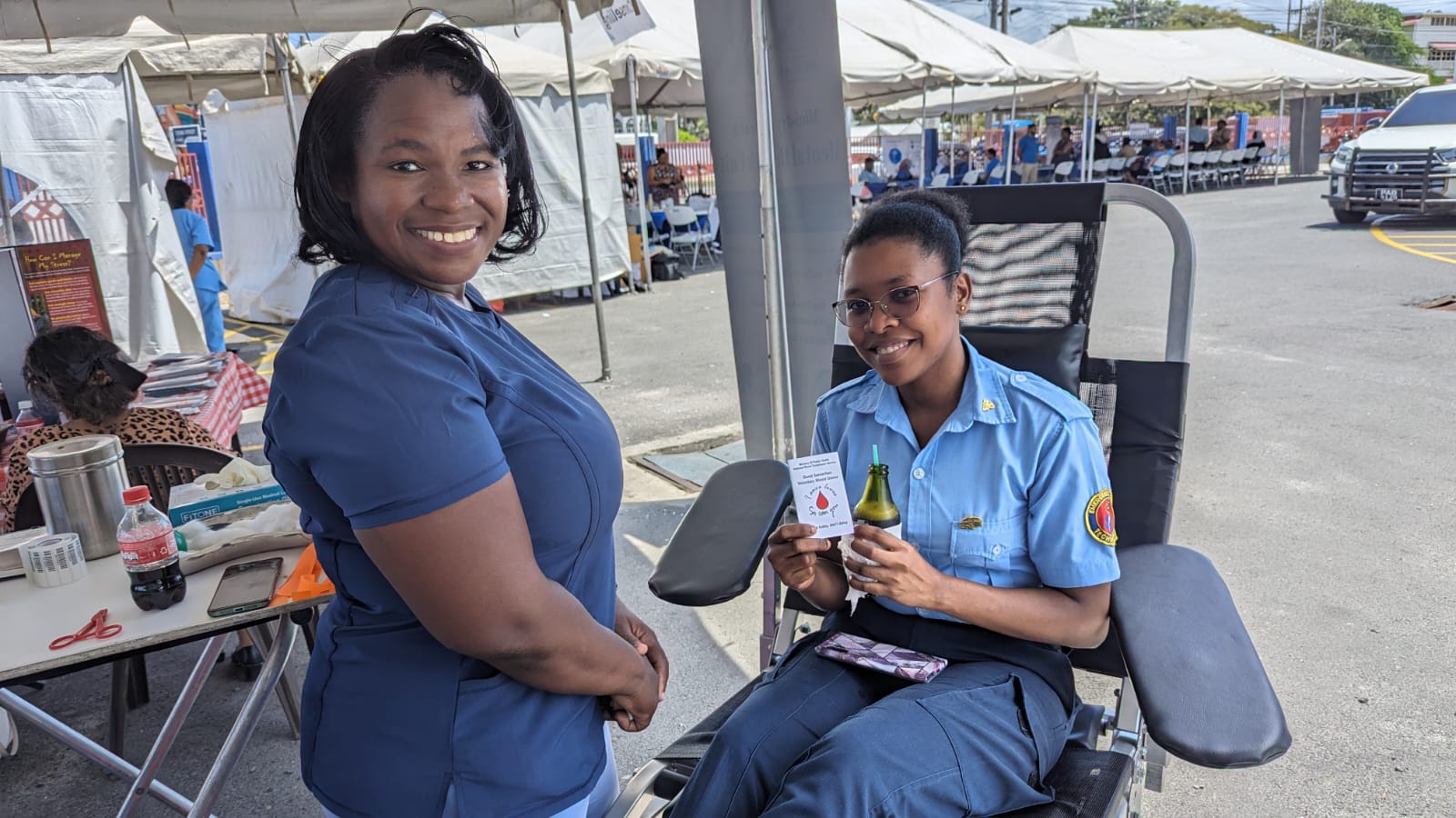 A women in blue medical scrub top stands while another women in collared uniform with a patch on the sleeve sits in a medical chair and holds up a paper card with a drawing of a blood drop on it.