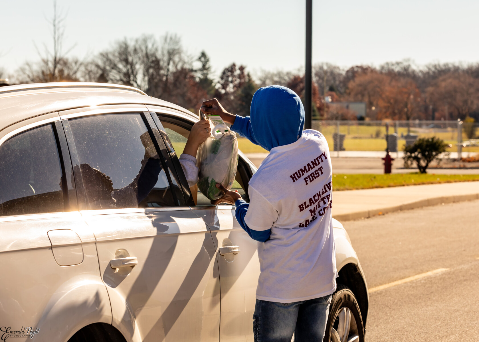 A person standing outside of a car gently hands a bag of food to an outstretched hand through the passenger window. The person outside of the car wears a white t shirt reading Humanity First and black lives matter on the back.