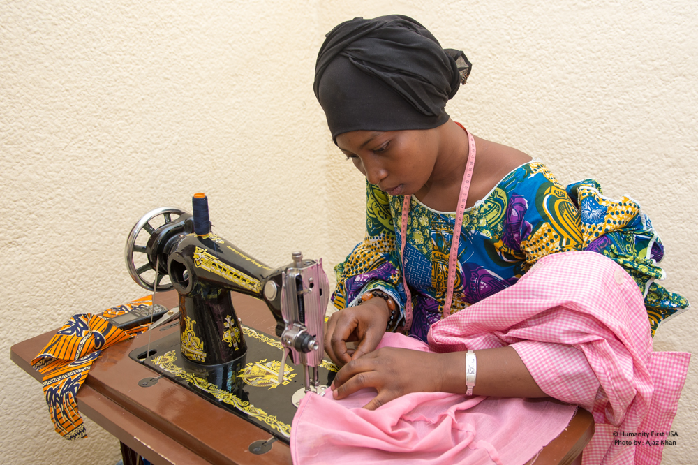 A woman in a bright patterned top and a black head wrap sews pink cloth on a black and gold sewing machine