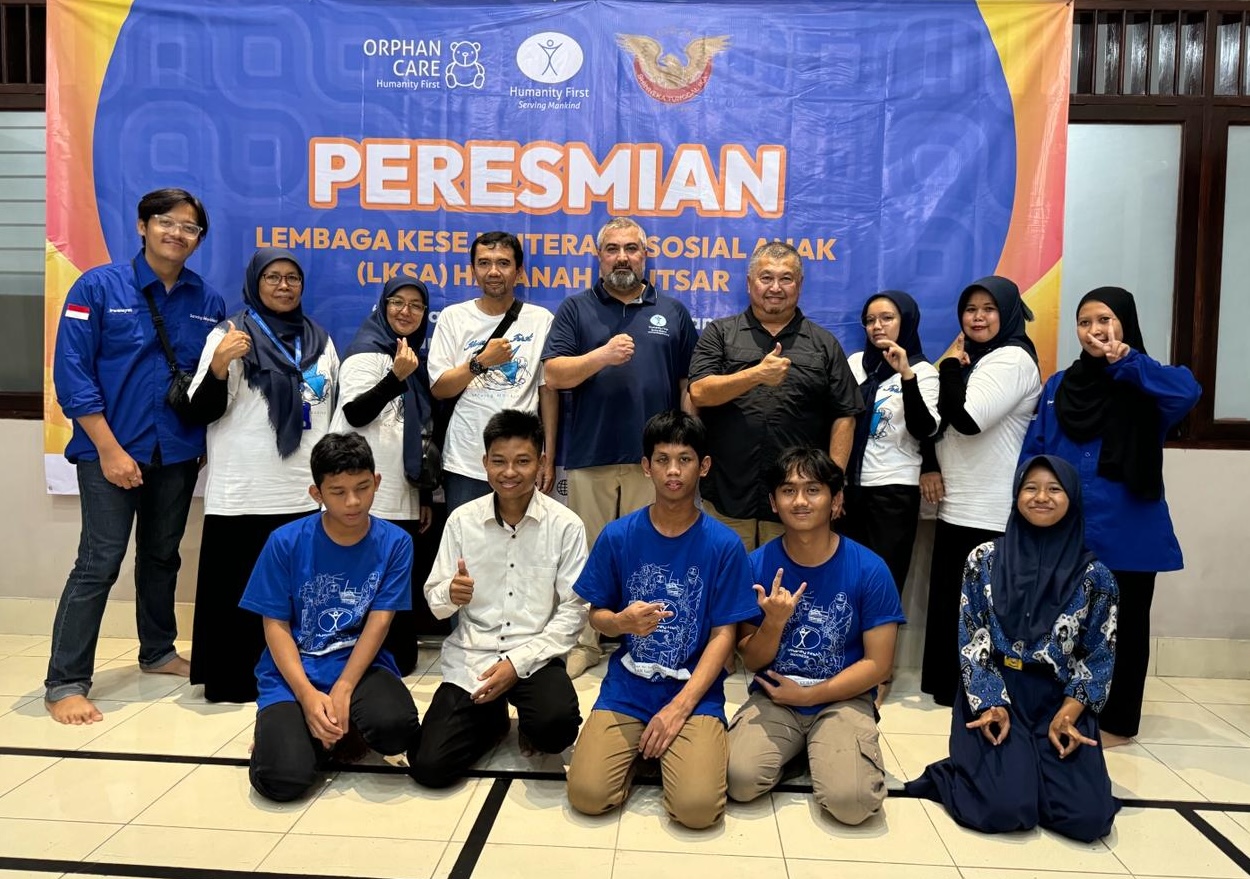 A mix of men, women, young men, and young women pose in two rows in front of a sign that says "Peresmian" with the logos of Humanity First and the orphan care program. Most of the people are making a thumbs up or a peace sign.