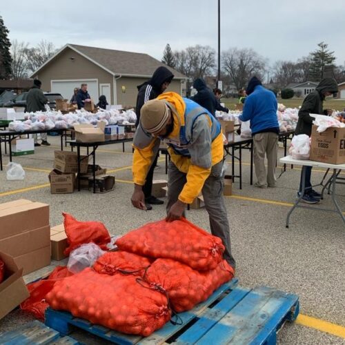 A man reaches for a large sack of potatoes while others in the background are packing plastic bags of food on long tables set up in a parking lot.