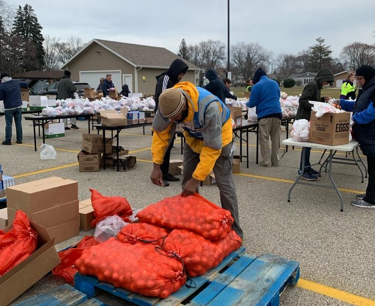 A man reaches for a large sack of potatoes while others in the background are packing plastic bags of food on long tables set up in a parking lot.