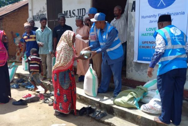 A man wearing a blue humanity first vest next hands a food package to a woman waering a long dress and shawl over her head. They stand outside a one story building on a dirt surface.