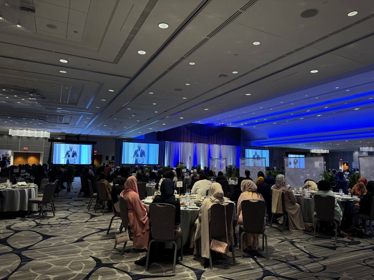 A view from the back of a large ballroom. Women in hijab sit at round dinner tables watching a speaker at the podium who is also projected onto digital screens.