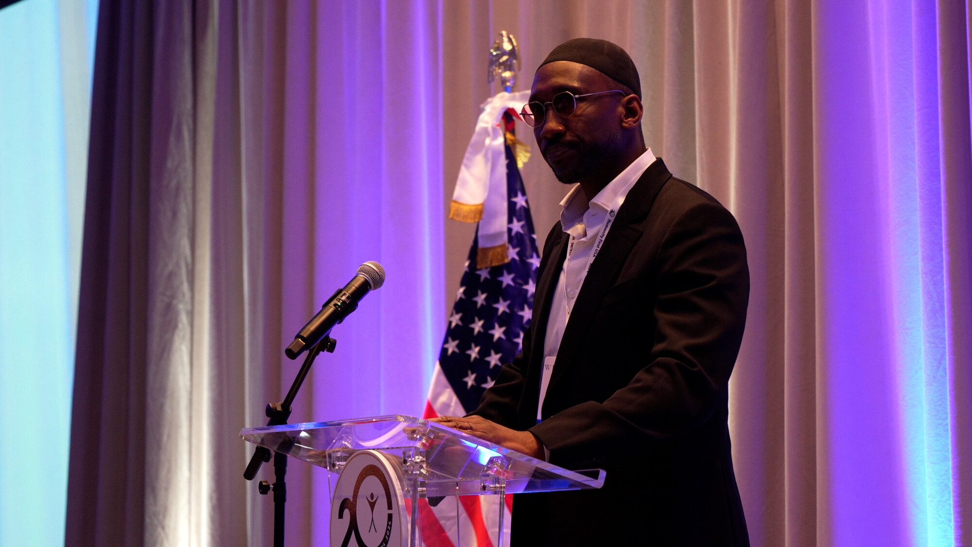 Mr. Mahershala Ali speaking from a podium with an American flag behind him