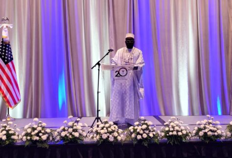 A man dressed in a white robe and cap of Western African style stands at the podium of a stage with white curtains behind it, white flowers in pots at the front edge, and an American flag on a stand to the viewer's left.