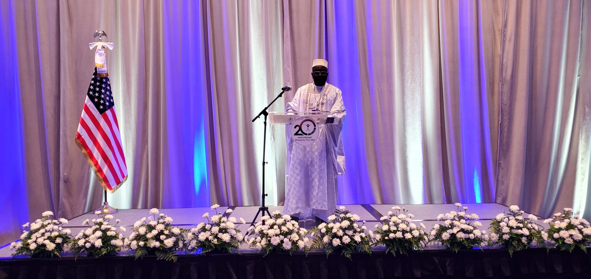 A man dressed in a white robe and cap of Western African style stands at the podium of a stage with white curtains behind it, white flowers in pots at the front edge, and an American flag on a stand to the viewer's left.
