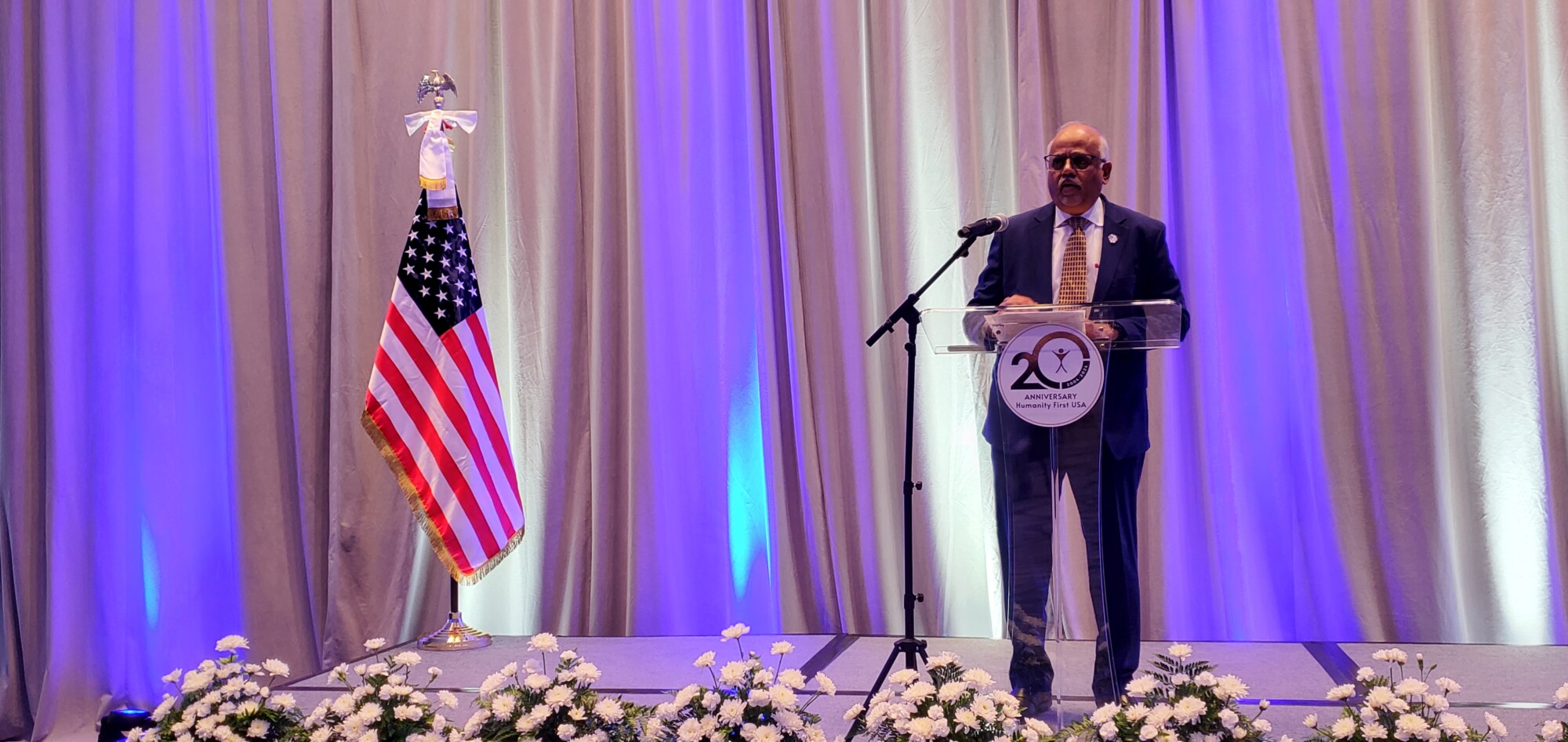 A man dressed in suit and tie stands at the podium of a stage with white curtains behind it, white flowers in pots at the front edge, and an American flag on a stand to the viewer's left.