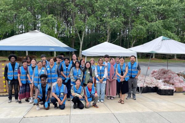 About two dozen people wearing blue humanity first vests over their street clothes pose in front of open air tents that have piles of bags and boxes of supplies underneath.